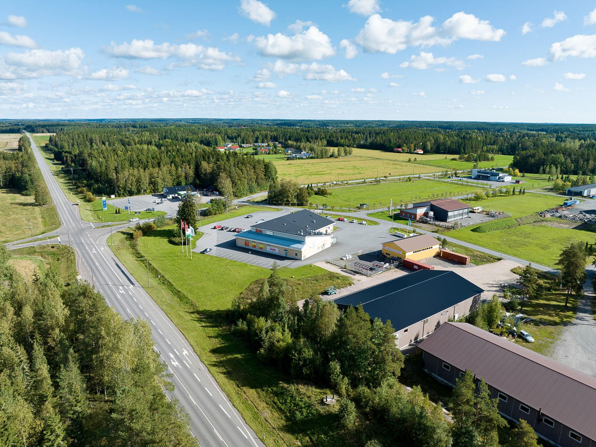 Ilmakuva maaseudun tienvarsikompleksista, jossa on rakennuksia, pysäköintialueita ja vihreitä peltoja sinisen taivaan alla.