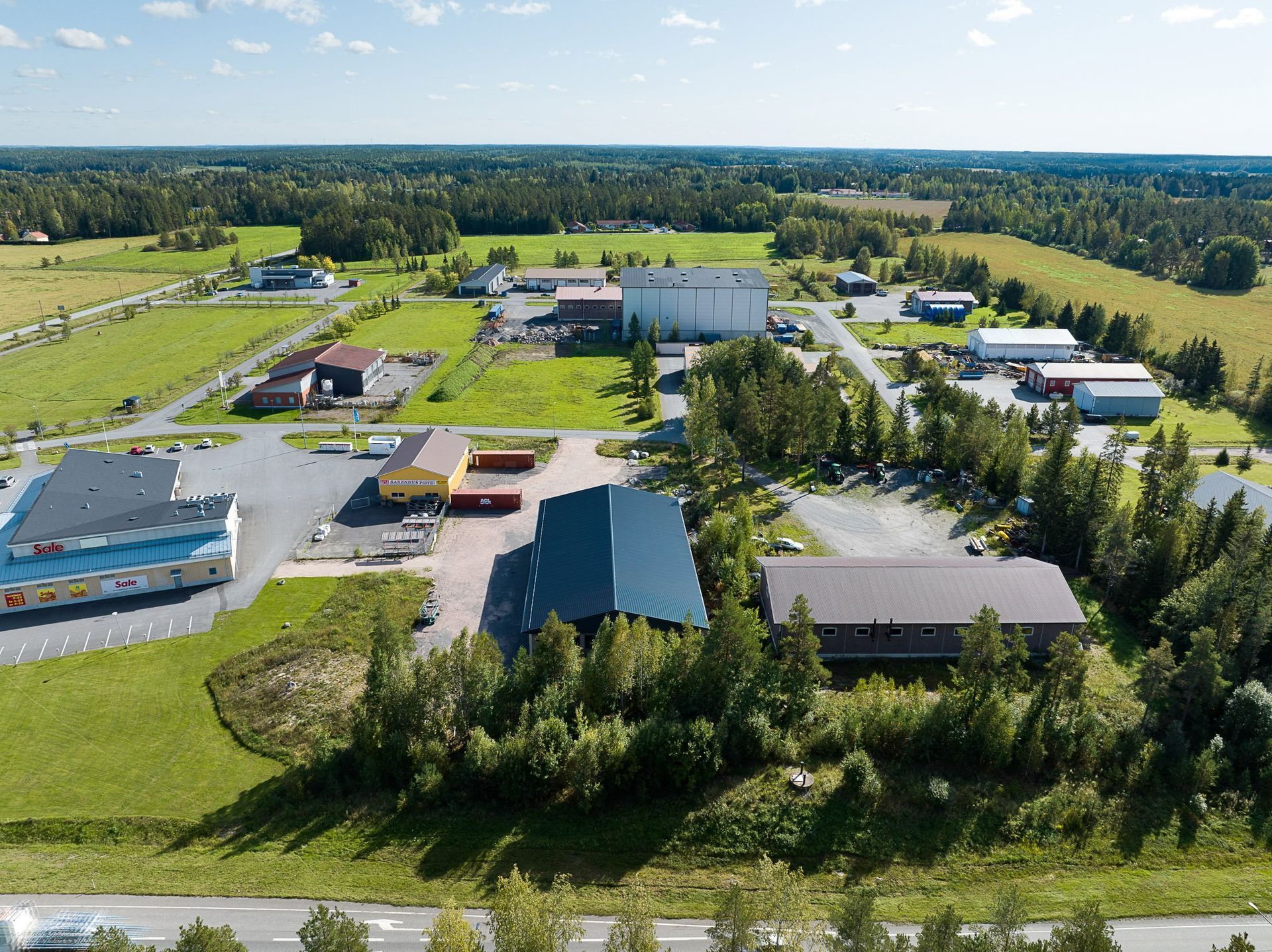 Ilmakuva maaseudun teollisuuspihasta, jossa on varastoja, kuorma-autoja ja puita kirkkaan taivaan alla