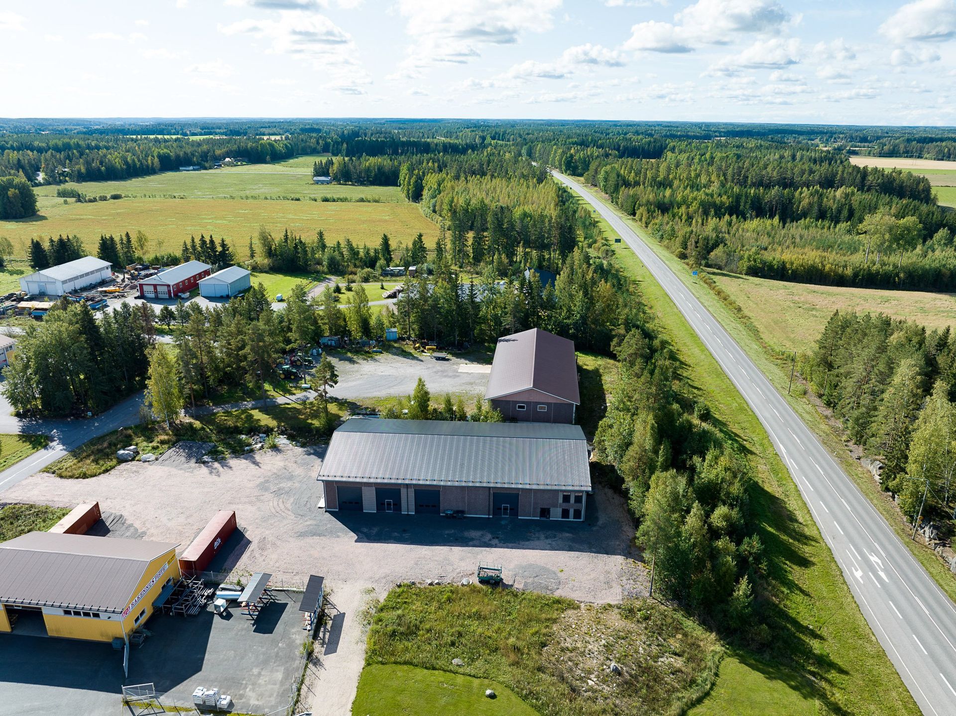 Ilmakuva maaseudun rakennuksista pitkän tien varrella, peltojen ja tiheän vihreän metsän ympäröimänä