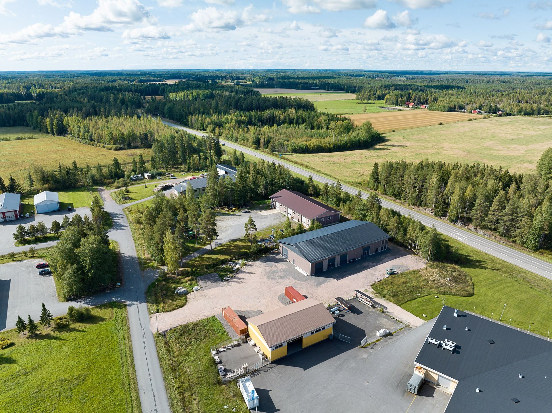 Ilmakuva maaseudun teollisuusalueesta, jossa on rakennuksia, teitä ja ympäröiviä vihreitä peltoja sinisen taivaan alla.
