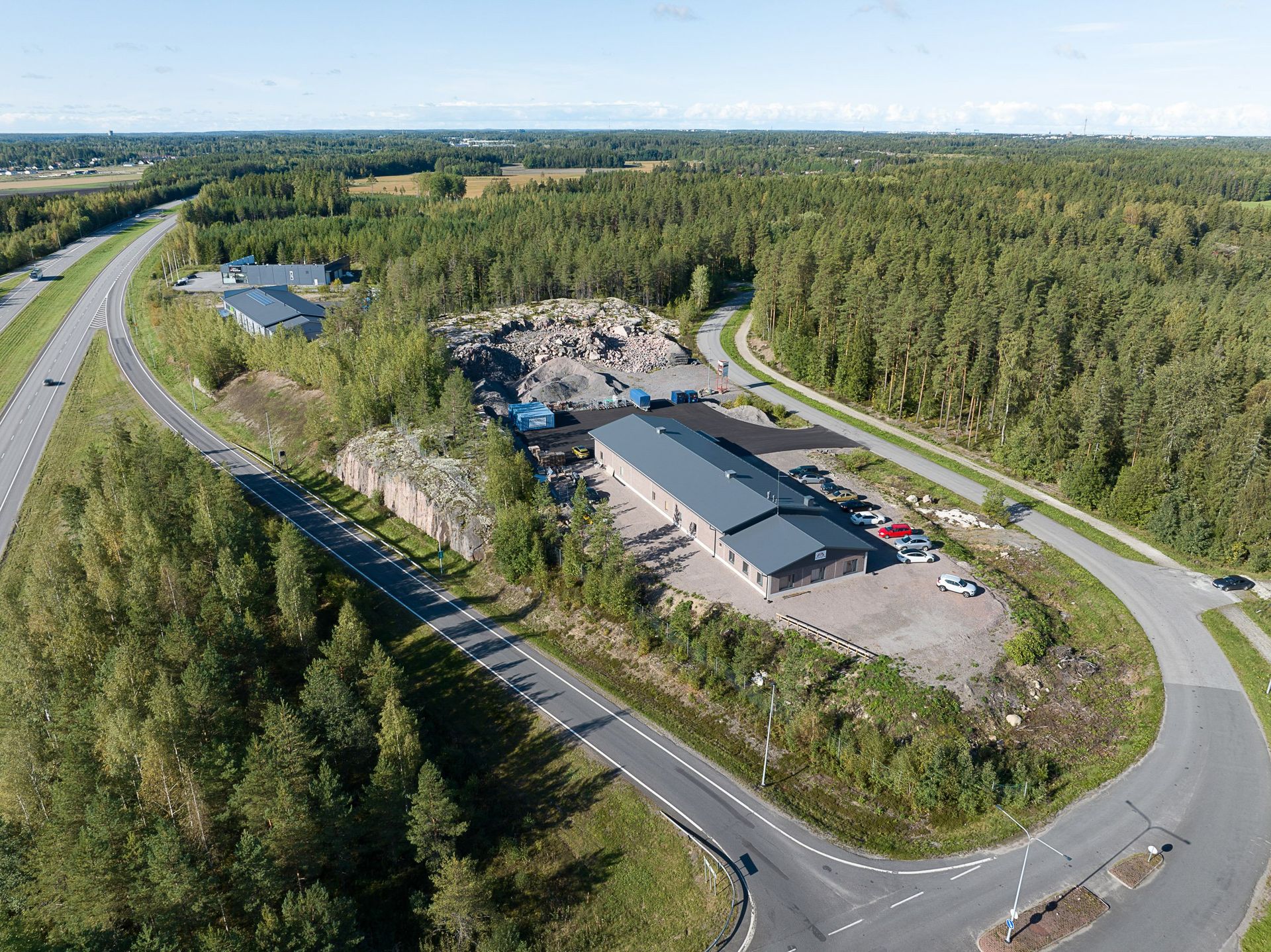 Ilmakuva liikerakennuksesta teiden ja tiheän metsän vieressä kirkkaan taivaan alla