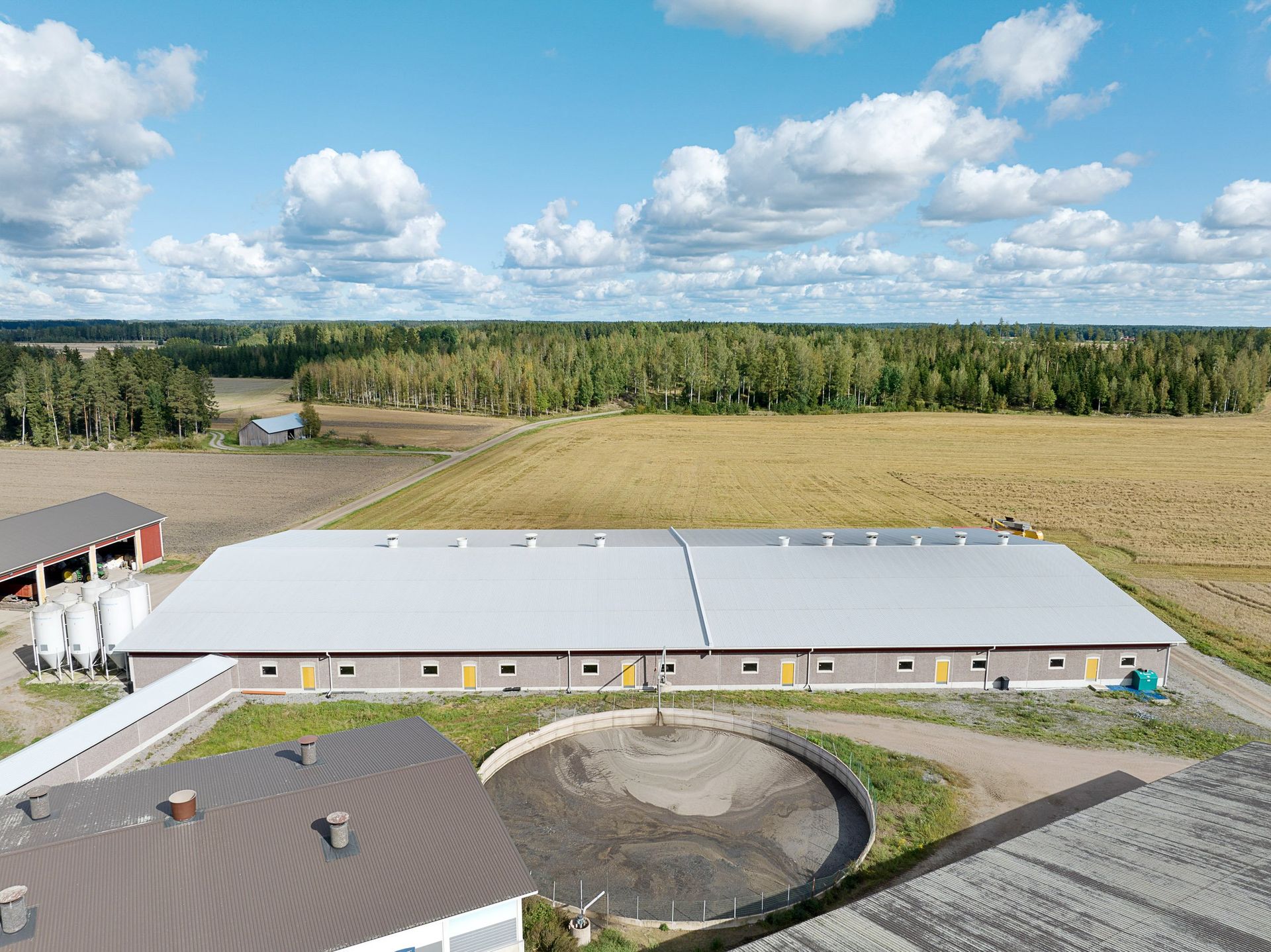 Ilmakuva pitkästä valkokattoisesta rakennuksesta peltojen, puiden ja pilvisen sinisen taivaan vierestä.