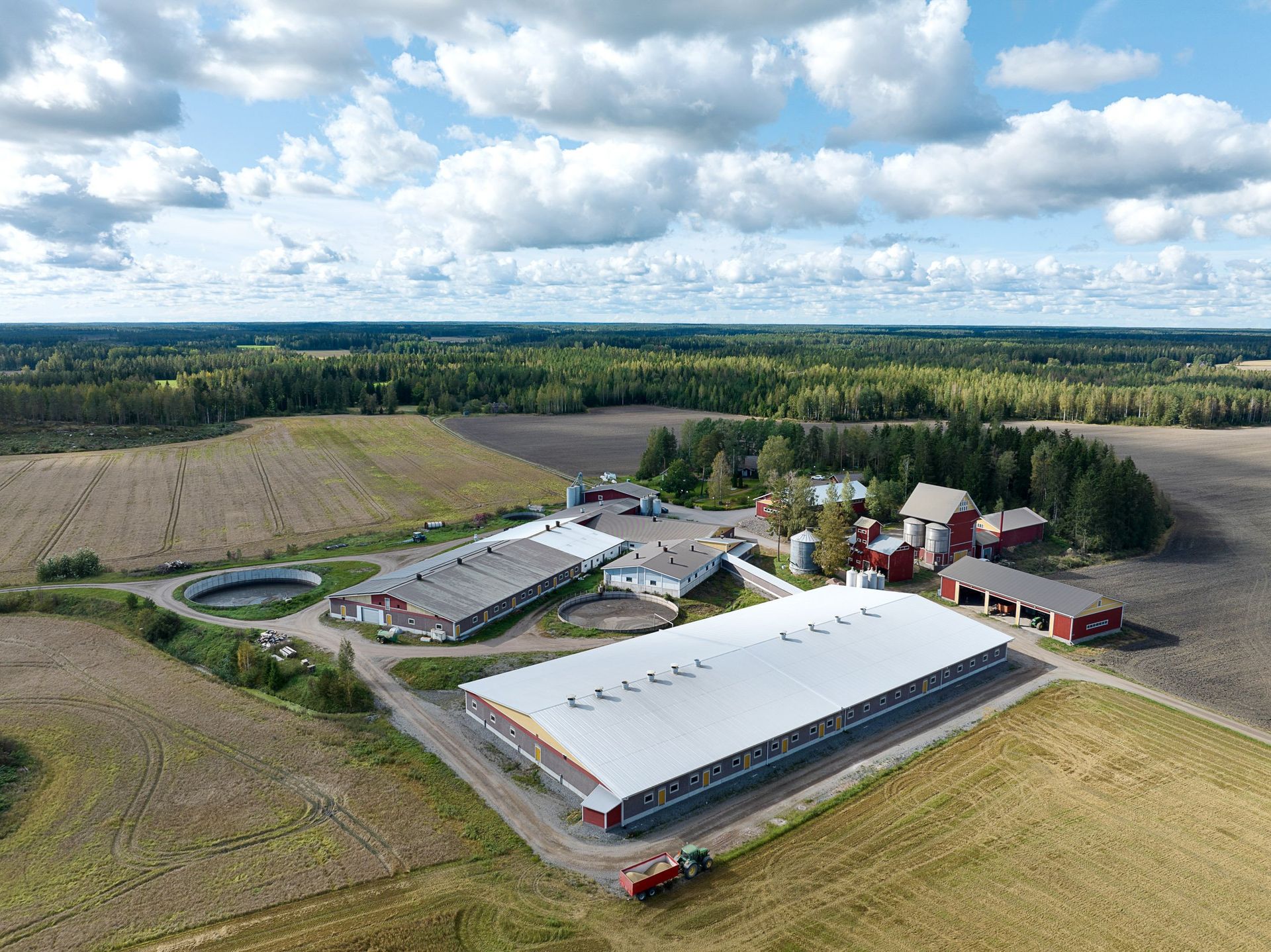 Ilmakuva suuresta maatilasta, jossa on valkokattoisia navettoja, peltoja ja puita pilvisen taivaan alla