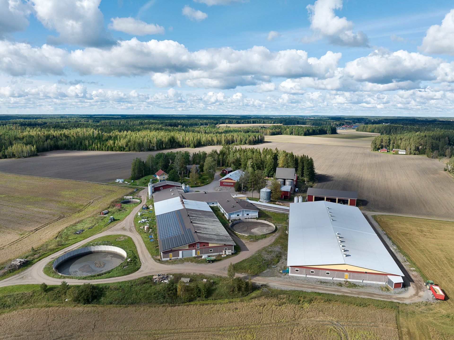 Ilmakuva maaseututilasta, jossa on navettoja, siiloja, peltoja ja lampi pilvisen taivaan alla.
