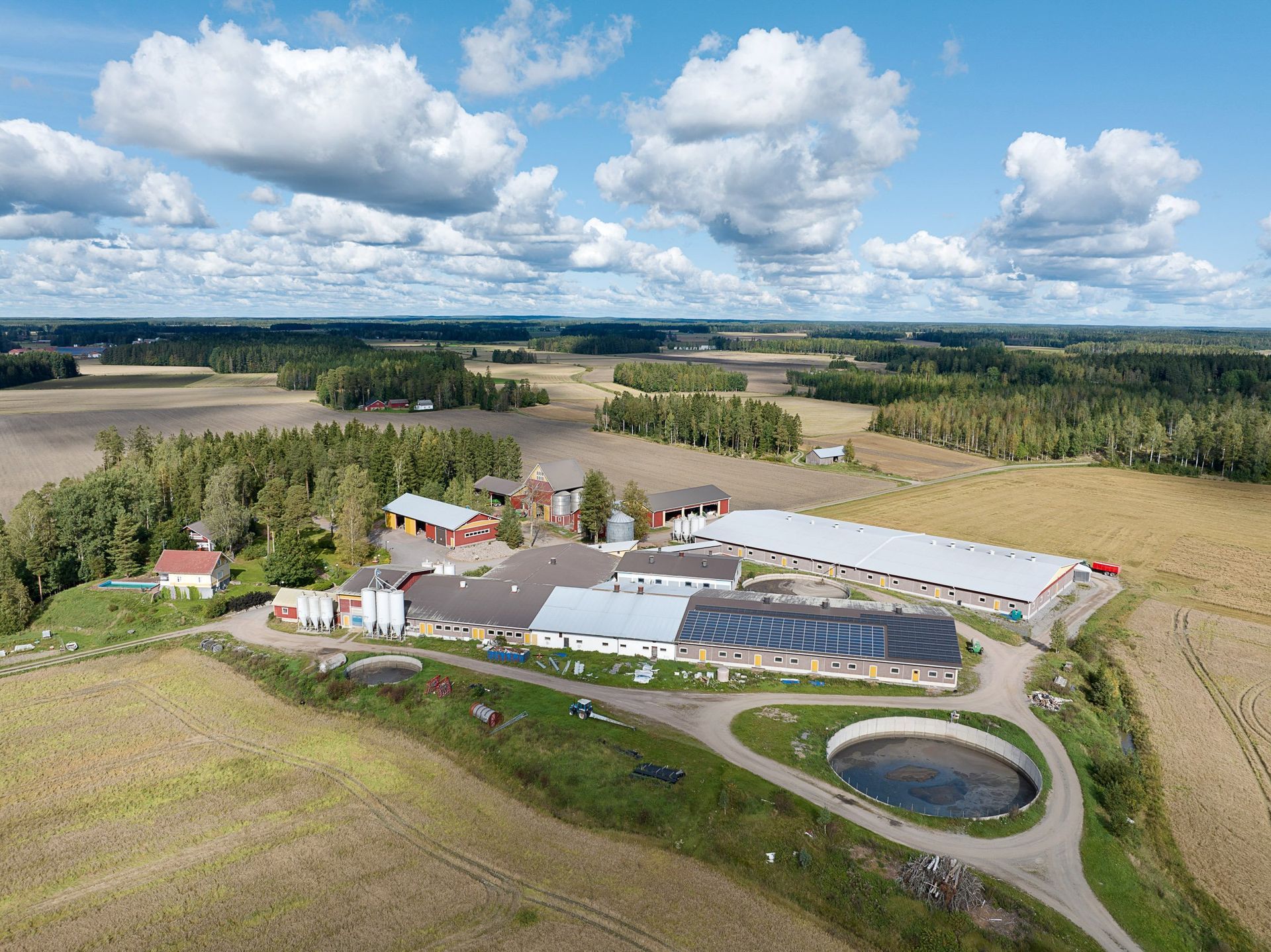 Ilmakuva maaseudun maatilakompleksista, jossa on navettoja, peltoja, puita ja pieni lampi pilvisen taivaan alla