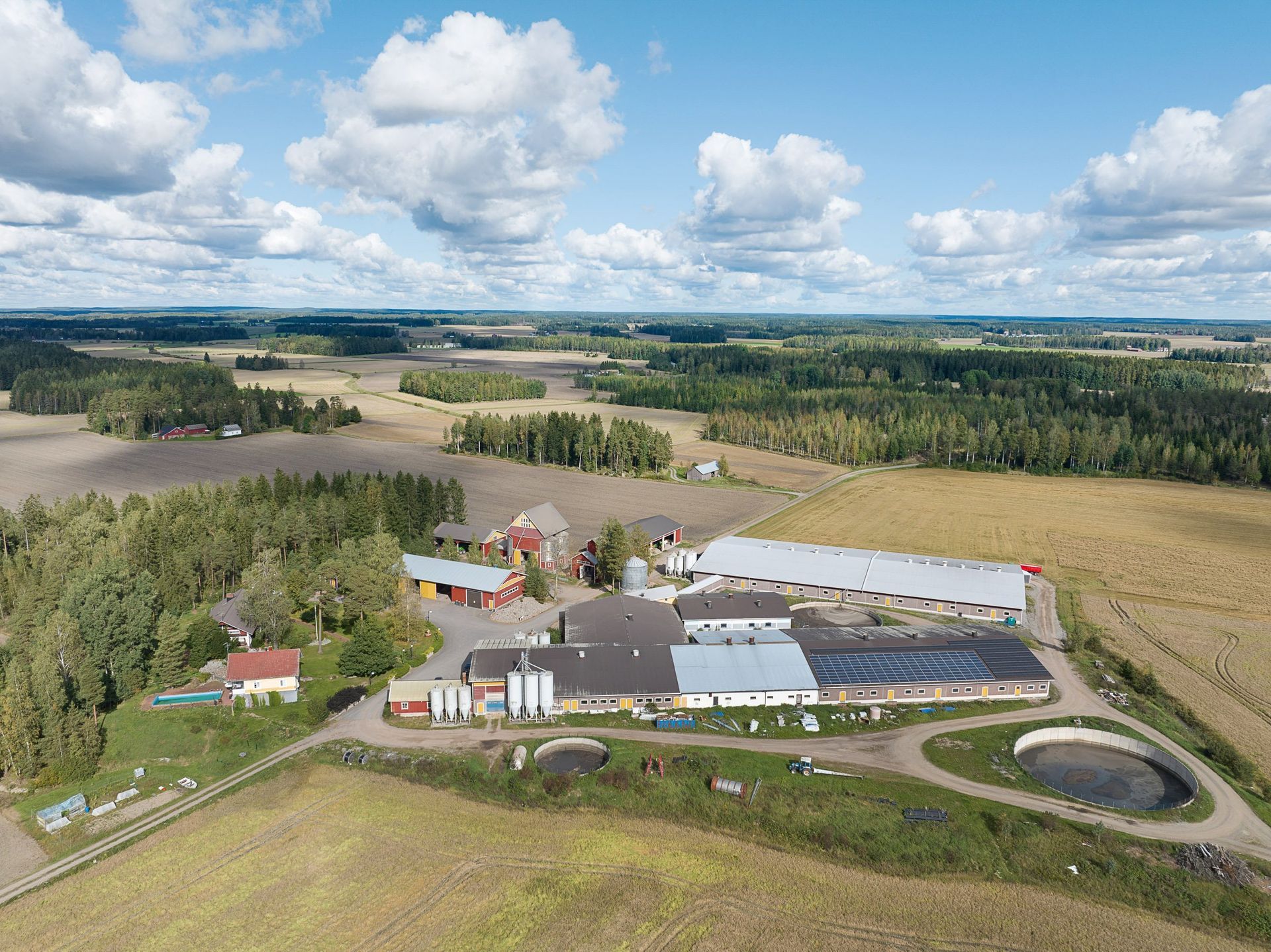Ilmakuva maatilasta, jossa on siiloja, navettoja, peltoja ja pyöreä lampi pilvisen sinisen taivaan alla