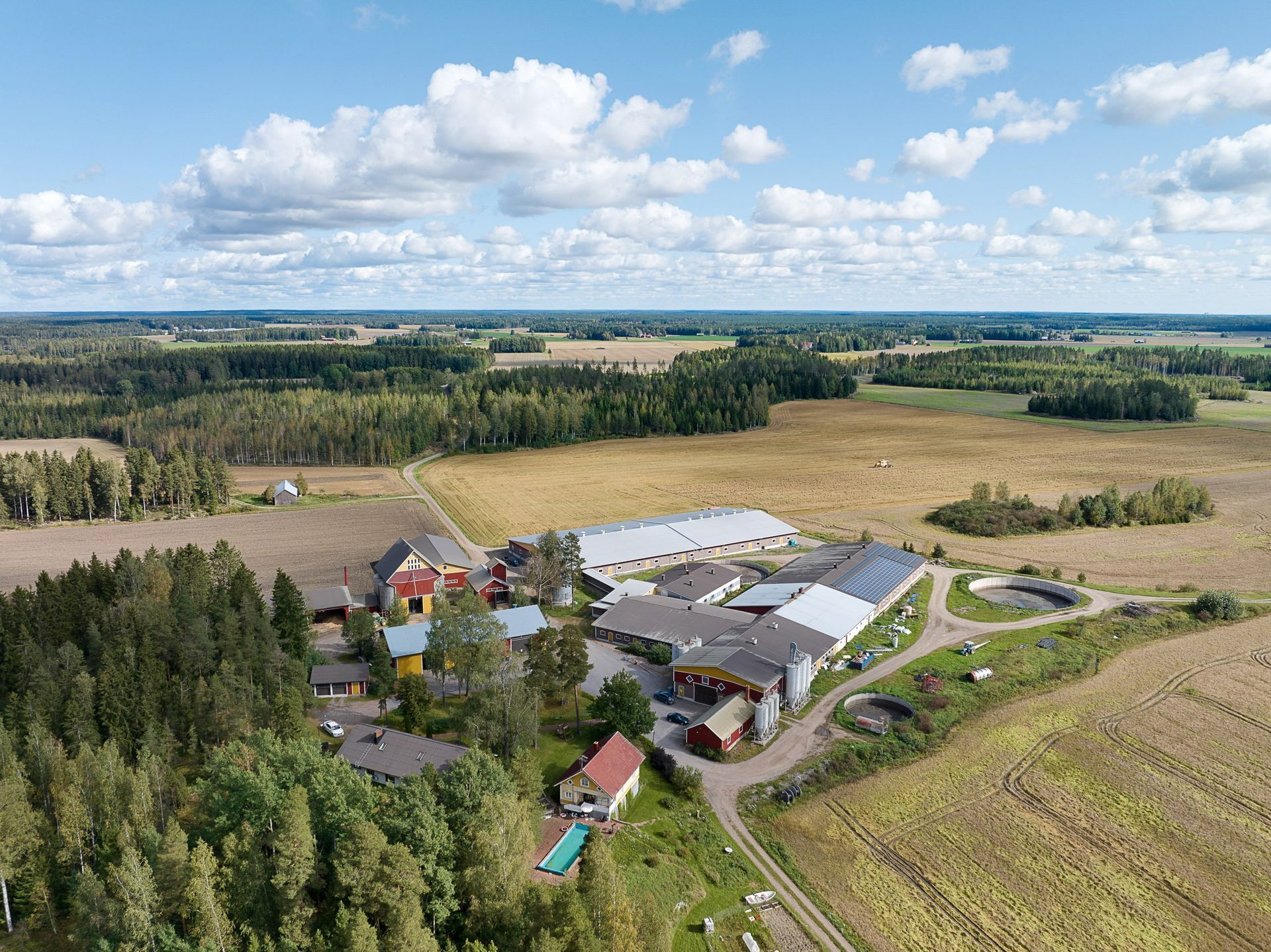 Ilmakuva maatilasta, jossa on peltoja, puita ja pilvinen taivas.