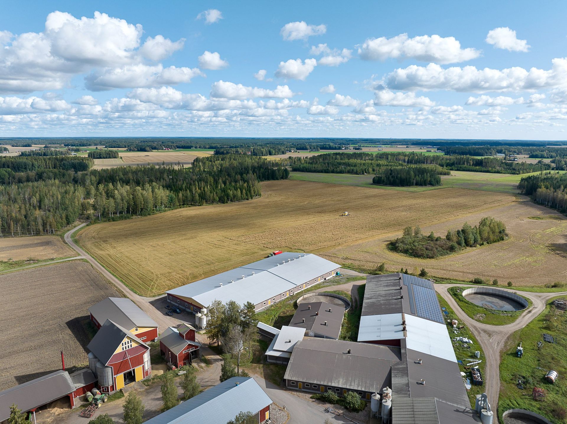 Ilmakuva maatilalta, jossa on navettoja, peltoja, puita ja pilvinen sininen taivas