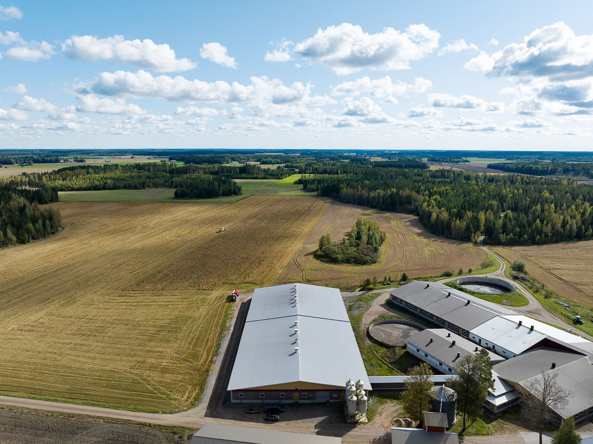 Ilmakuva maatilan rakennuksista peltojen ja metsän vieressä pilvisen sinisen taivaan alla