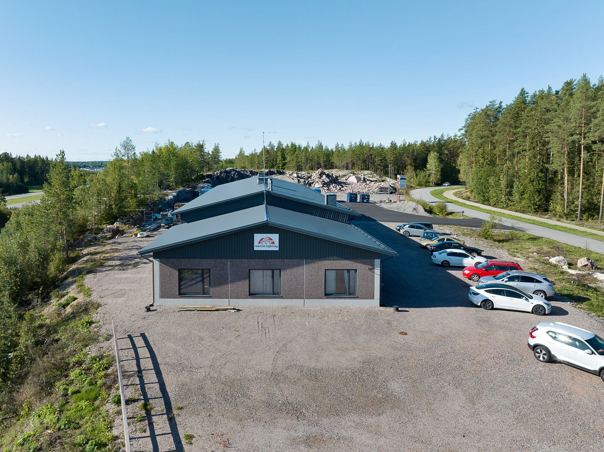Ilmakuva suuresta tummakattoisesta rakennuksesta, jossa on pysäköityjä autoja puiden reunustaman tien varrella.