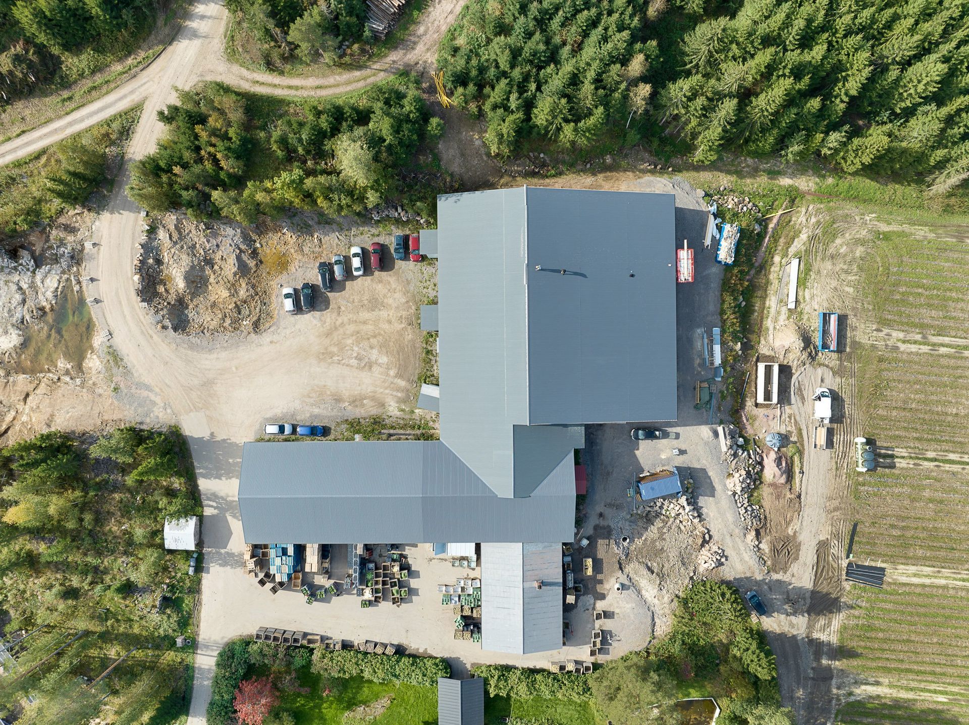 Ilmakuva maaseudun talosta, jossa on harmaa katto, sorapäällysteinen ajotie, pysäköityjä ajoneuvoja ja ympäröiviä puita.
