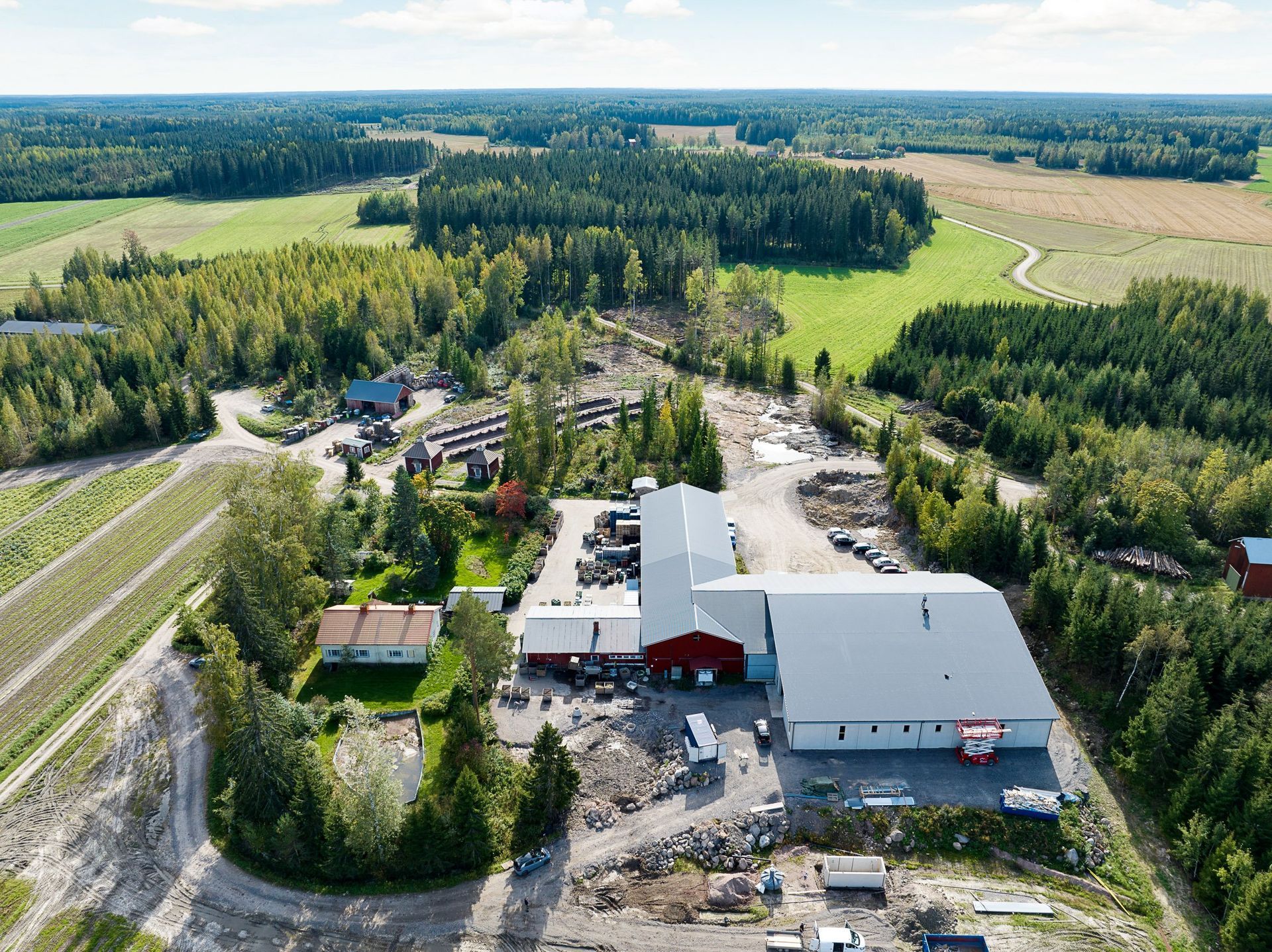 Ilmakuva maaseudun maatilakompleksista, jossa on punavalkoisia rakennuksia, peltoja ja ympäröivää metsää.