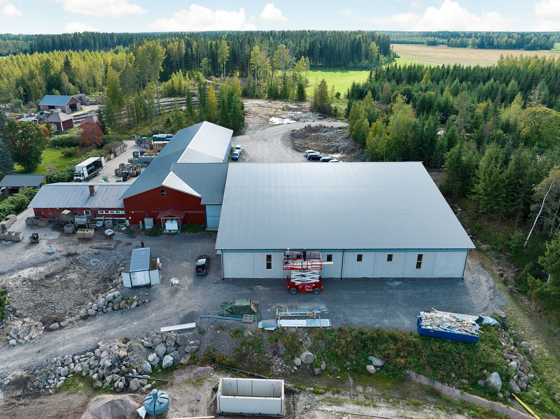 Ilmakuva maaseudun maatilakompleksista, jossa on punavalkoisia rakennuksia puiden ja sorapihojen ympäröimänä.