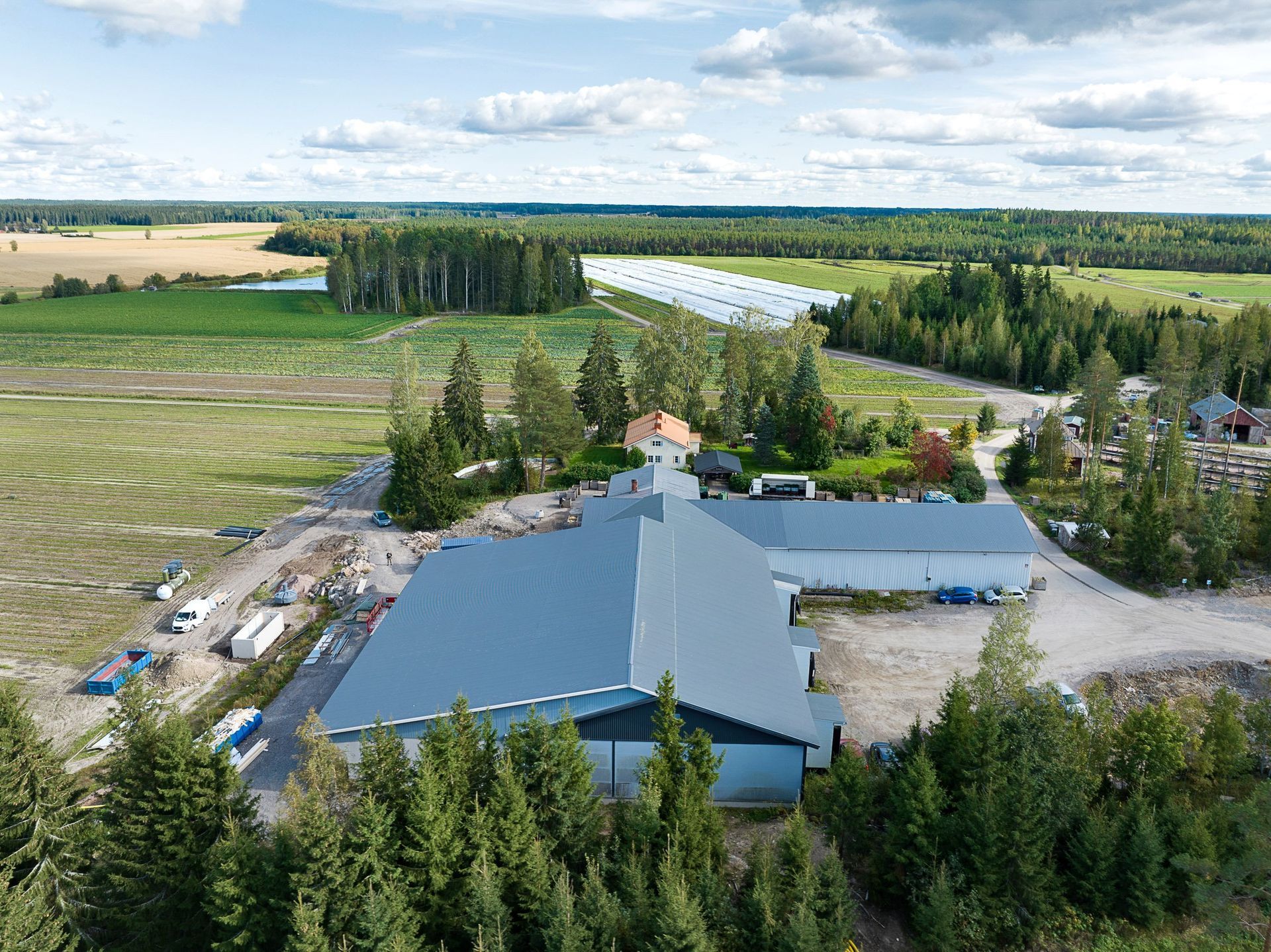 Ilmakuva maaseudun maatilarakennuksesta, jossa on sininen katto, puiden, peltojen ja mutkittelevan joen ympäröimänä.