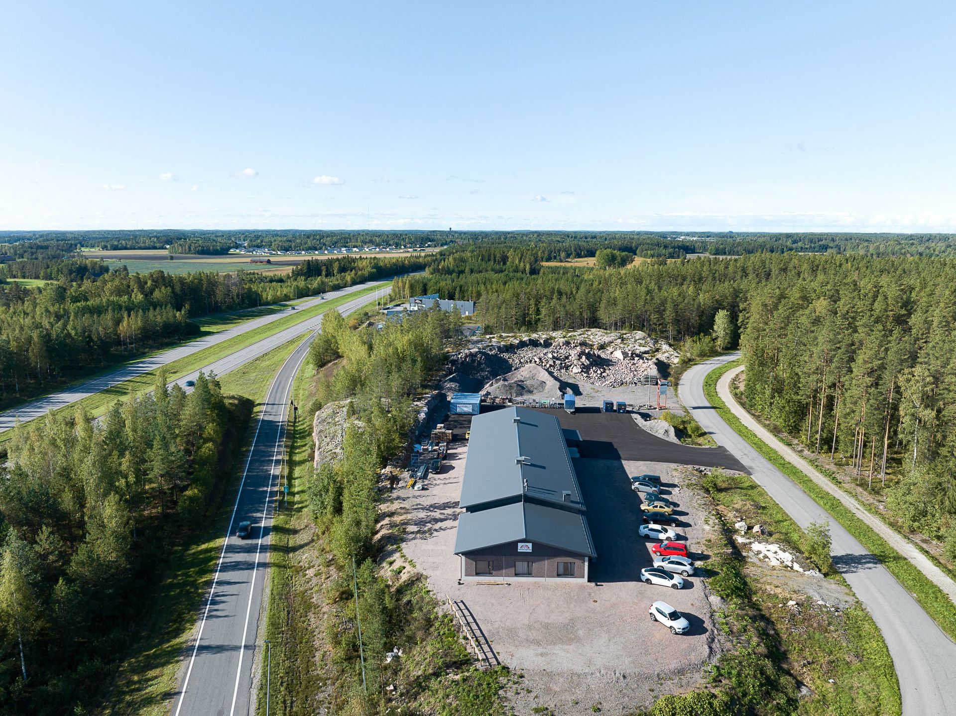 Ilmakuva pitkästä rakennuksesta ja pysäköintialueesta moottoritien vieressä, puiden ympäröimänä kirkkaan taivaan alla