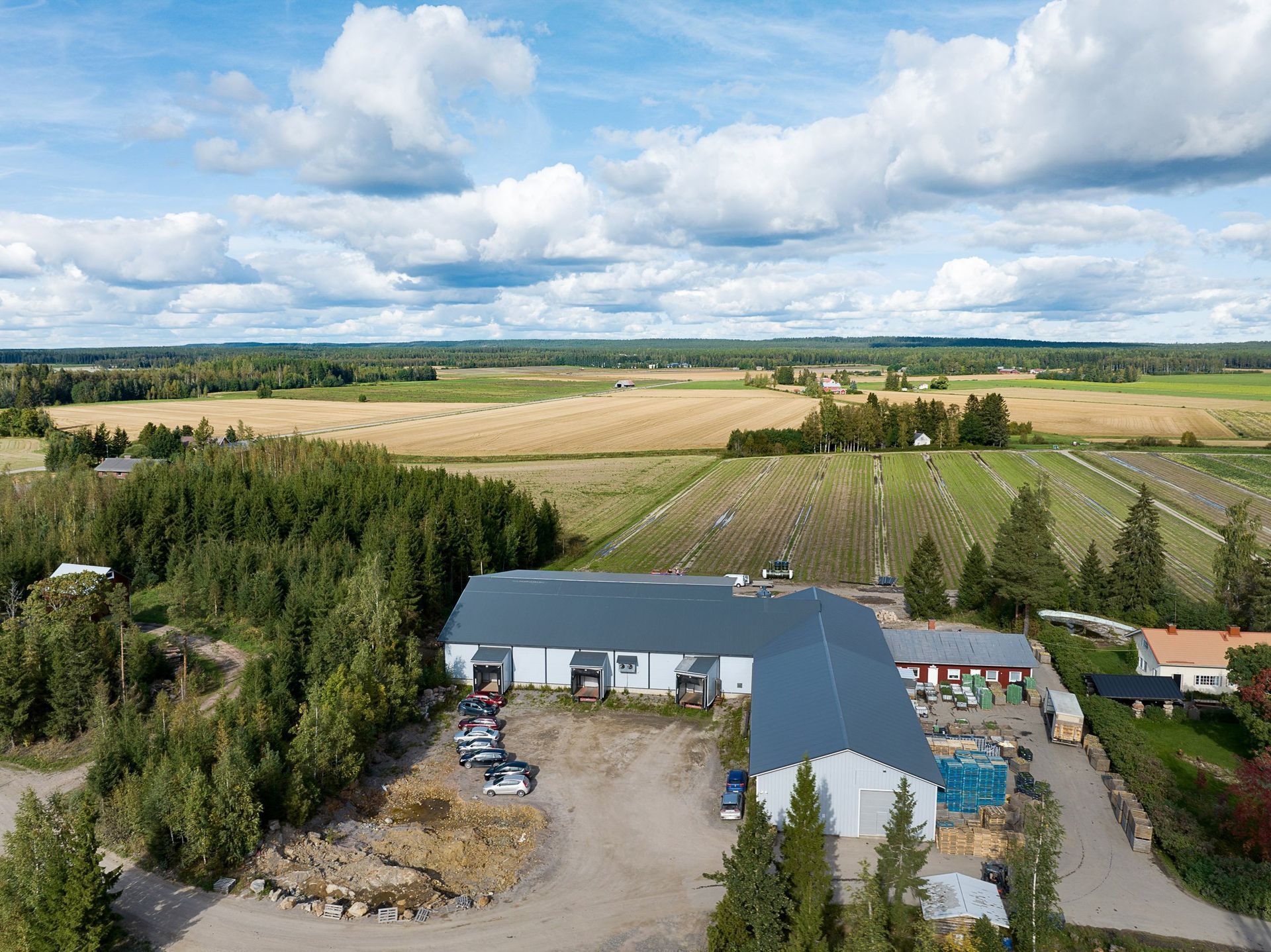 Ilmakuva maatilan pihasta rakennuksineen ja koneineen, peltojen ja puiden ympäröimänä pilvisen taivaan alla