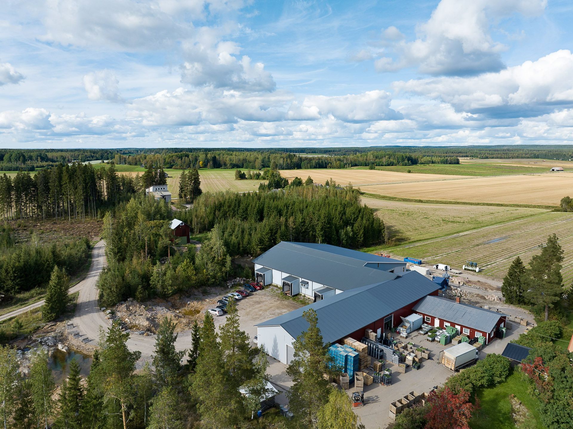Ilmakuva maaseudun maatilalta, jossa on harmaakattoisia rakennuksia, puita, peltoja ja osittain pilvinen taivas.
