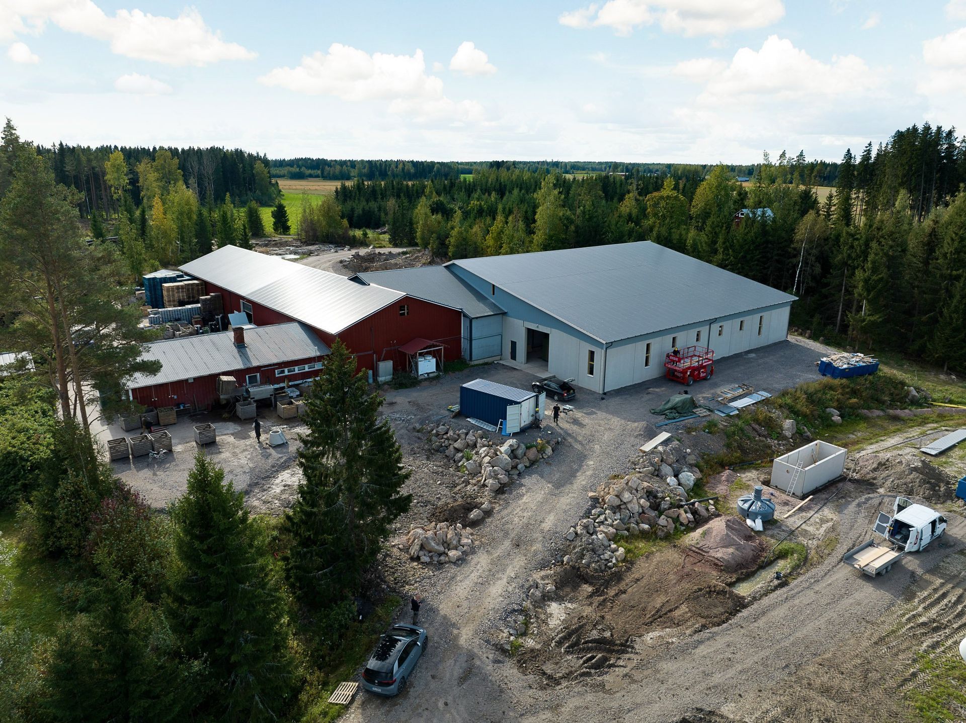 Ilmakuva maaseudun maatilasta, jossa on punaisia ​​ja harmaita navettoja, kuorma-autoja sekä ympäröiviä puita ja hiekkateitä