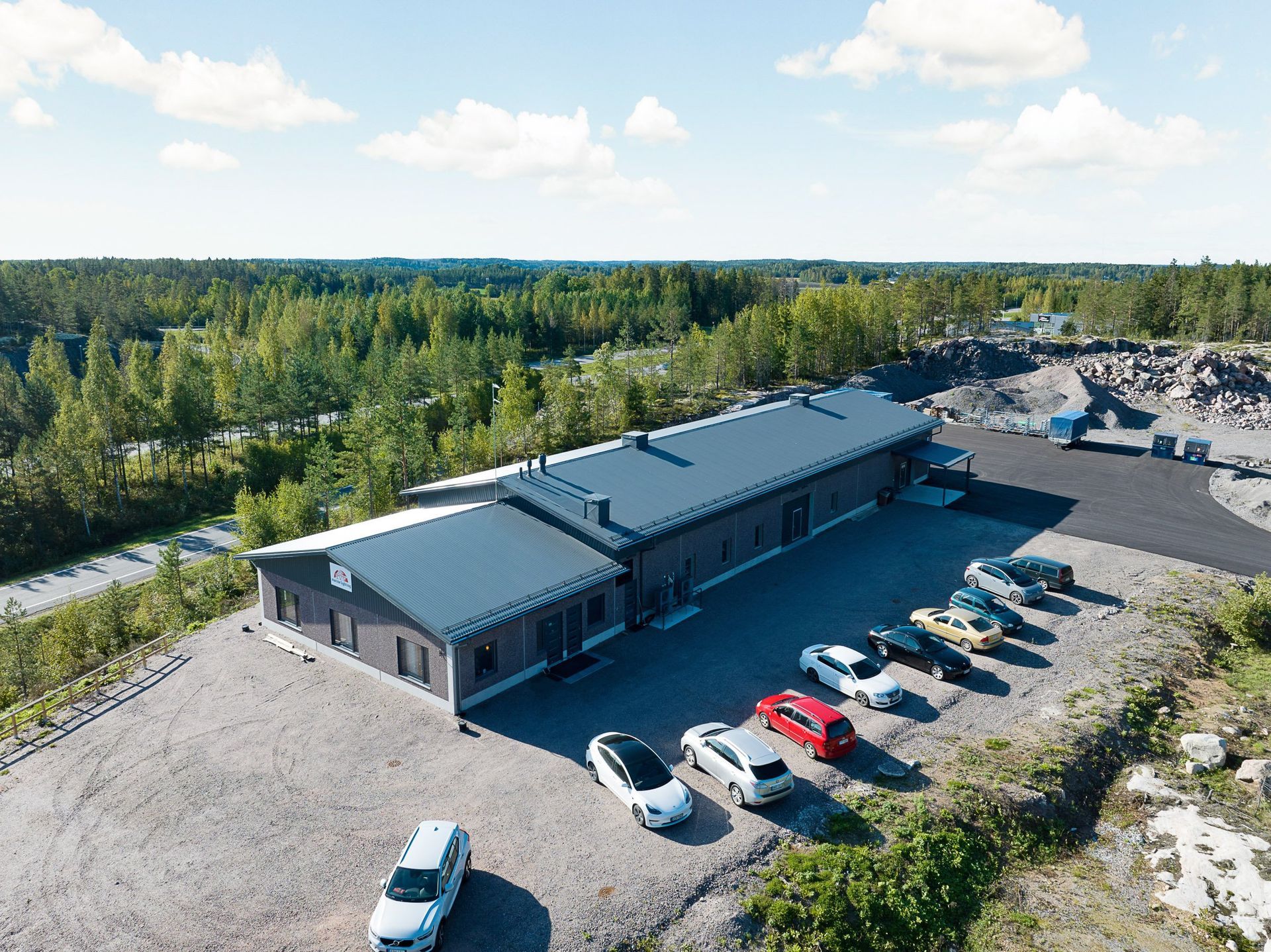 Ilmakuva pitkästä harmaasta rakennuksesta sorakentän vieressä, jossa on pysäköityjä autoja metsän ympäröimänä.
