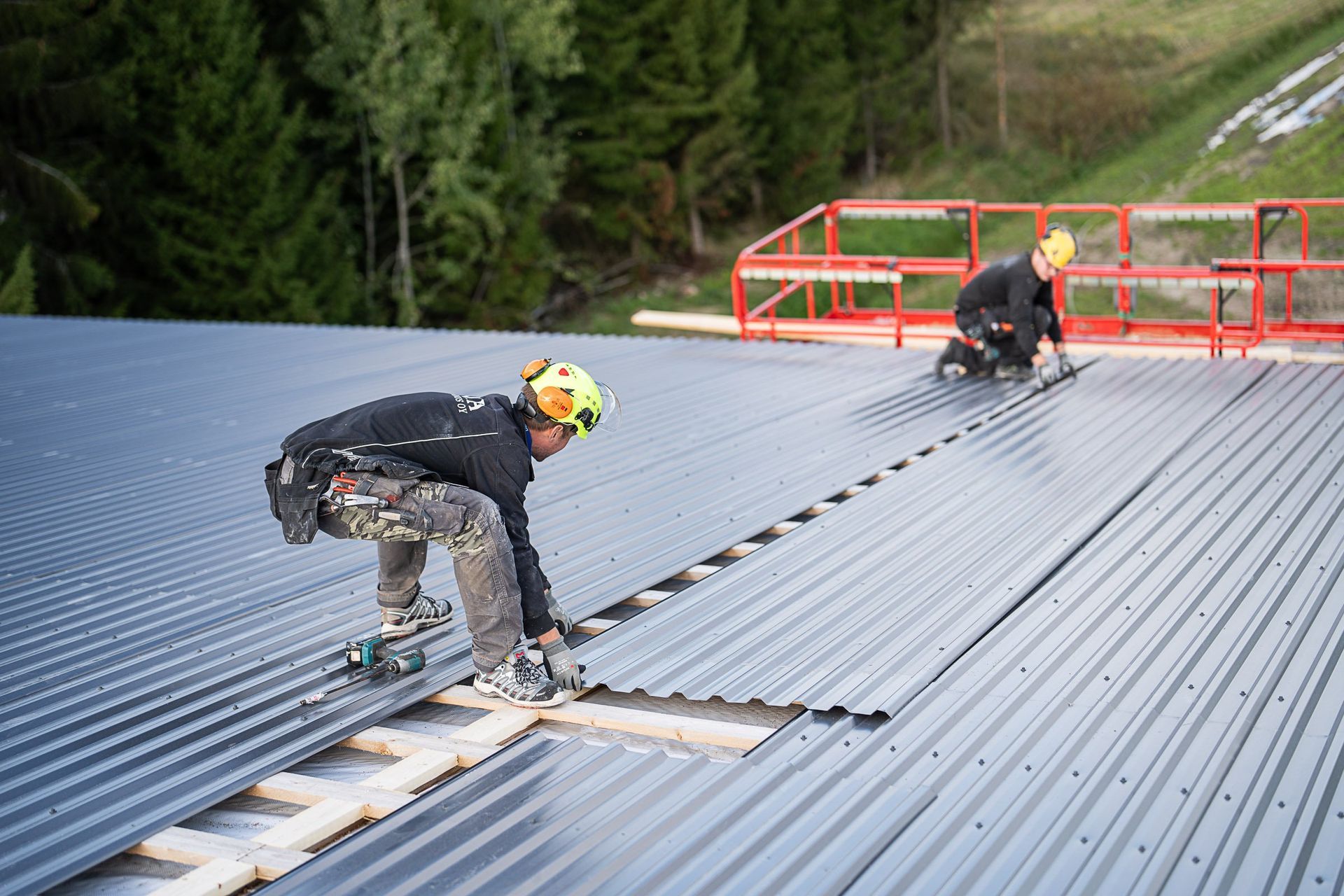 Kaksi työntekijää asentamassa aaltopeltikattoa kaltevalle katolle metsäisen rinteen lähellä