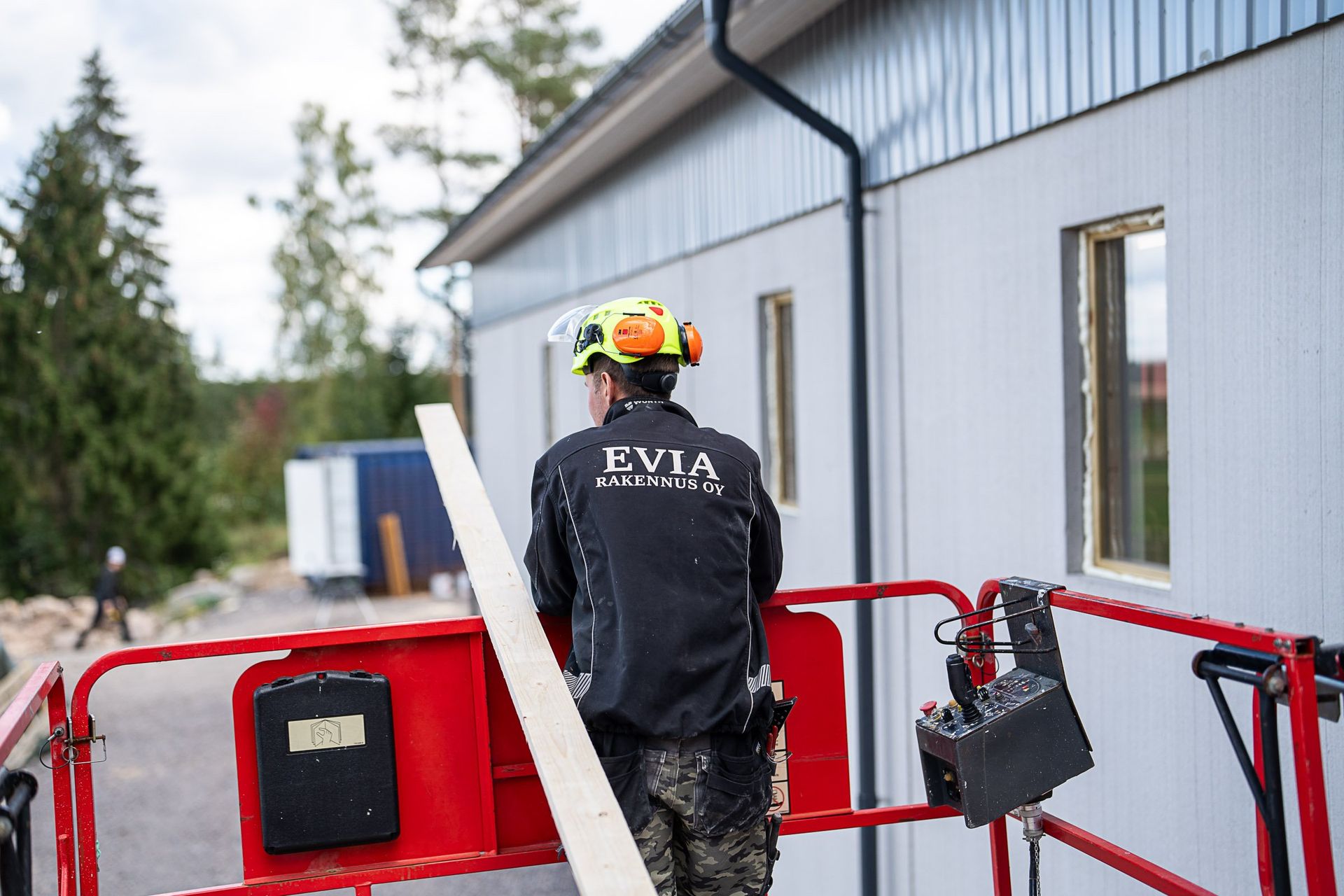 Kypäräpäinen työntekijä seisoo punaisessa hississä harmaan rakennuksen vieressä kantaen mustaa laukkua.