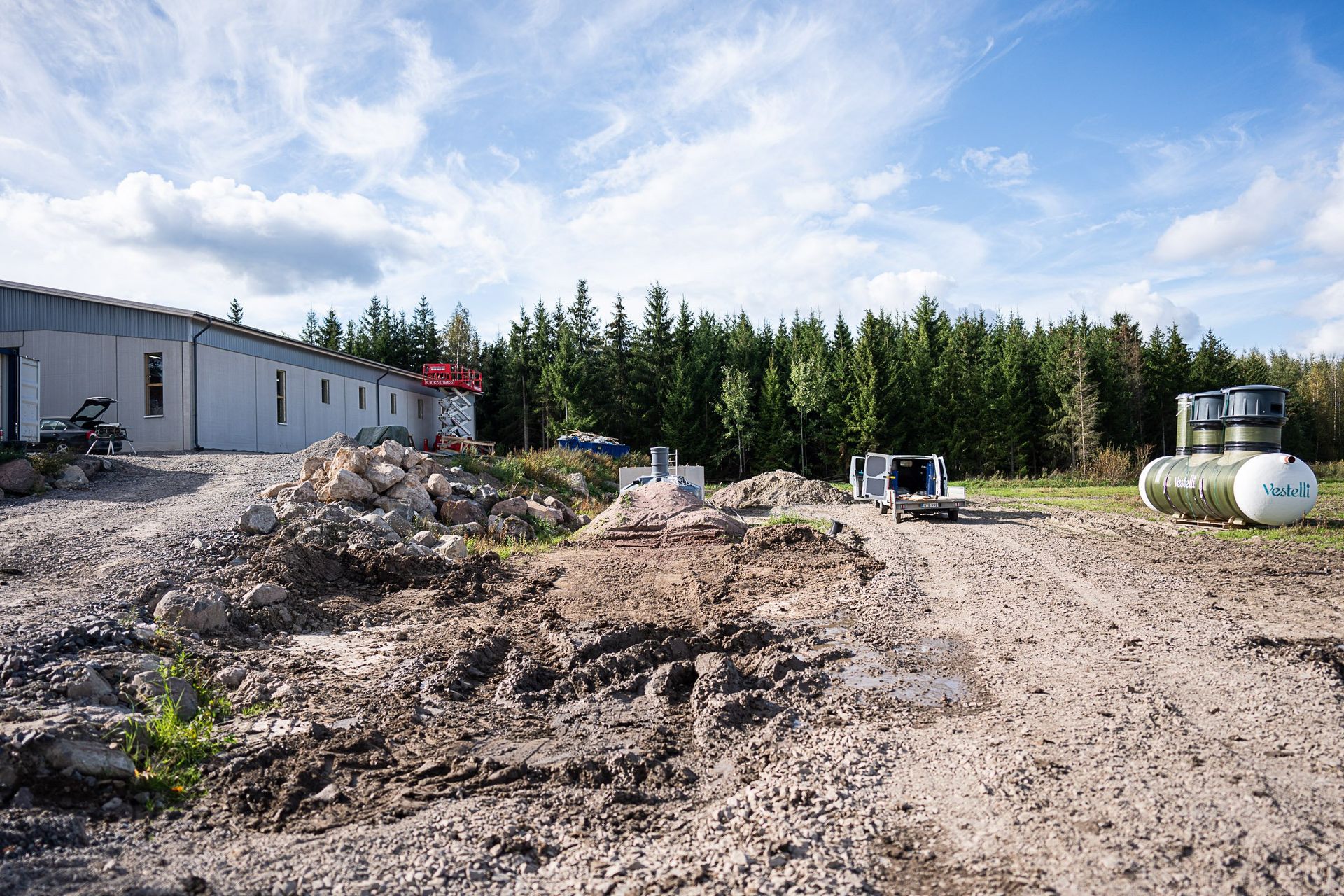 Rakennustyömaa, jossa on maakasoja metallirakennuksen vieressä, puita ja yleishyödyllisiä säiliöitä pilvisen taivaan alla
