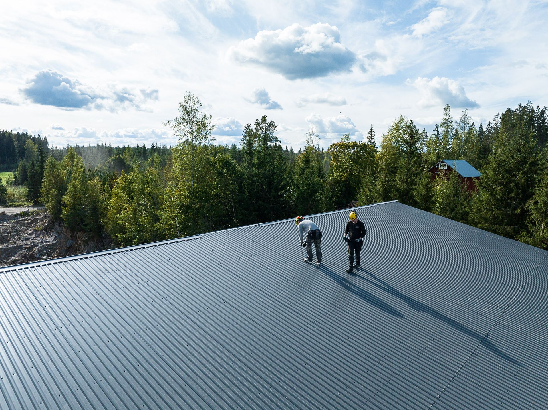 Kaksi retkeilijää seisoo aaltopeltikatolla metsämaiseman yllä pilvisen taivaan alla