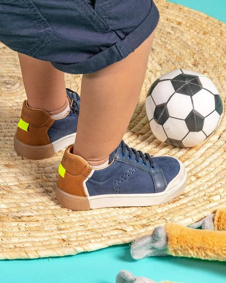 Pieds d'enfant chaussés de baskets bleues sur un tapis de paille, à côté d'un ballon de football et d'un poisson en plastique, sur fond turquoise.