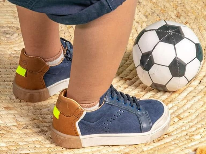 Pieds d'enfant chaussés de baskets bleues sur un tapis tressé, à côté d'un ballon de football.