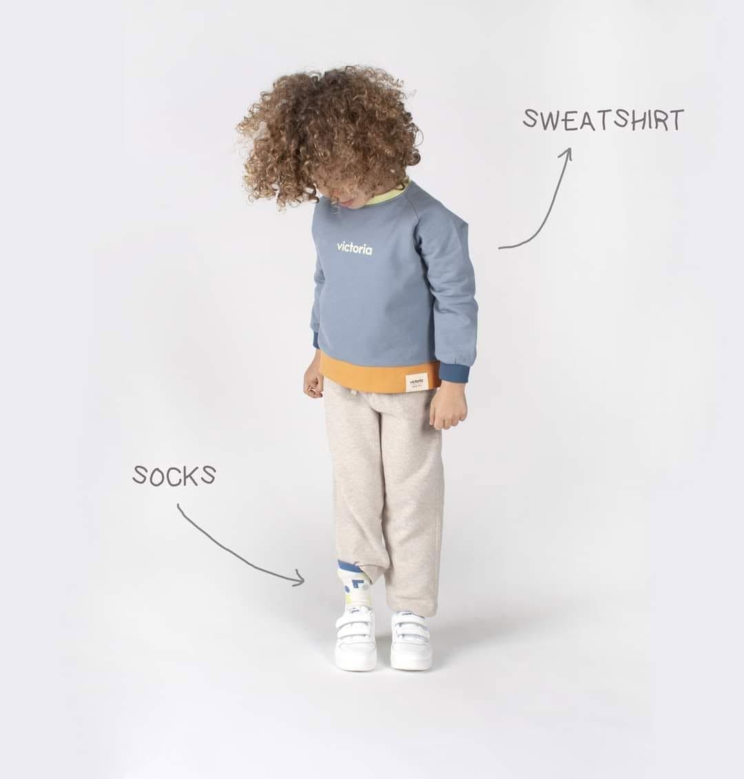 Enfant portant un sweat-shirt bleu, un pantalon beige et des chaussettes blanches sur fond blanc uni, avec des étiquettes indiquant les vêtements.