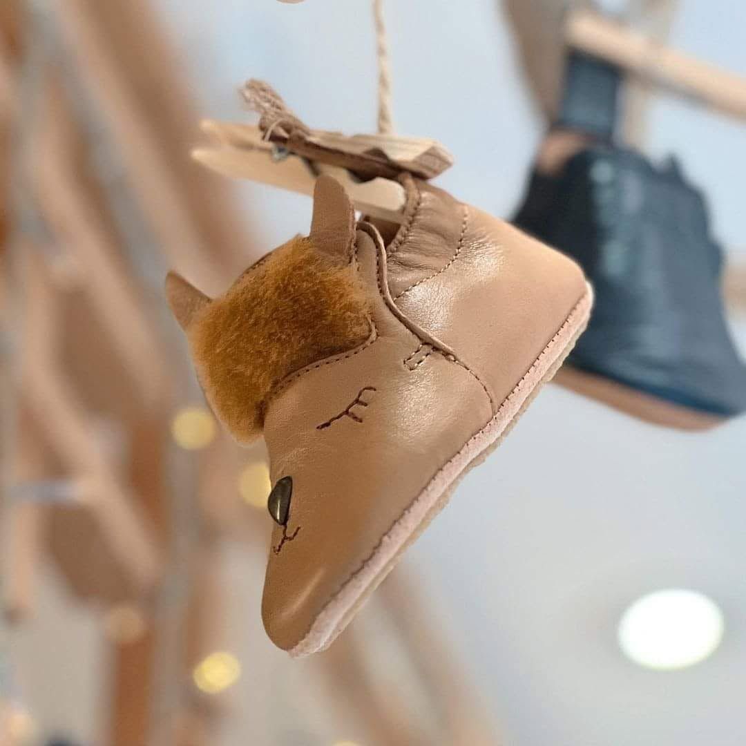 Chausson bébé en daim beige avec bordure en fourrure et surpiqûres, suspendu par une ficelle dans une vitrine.