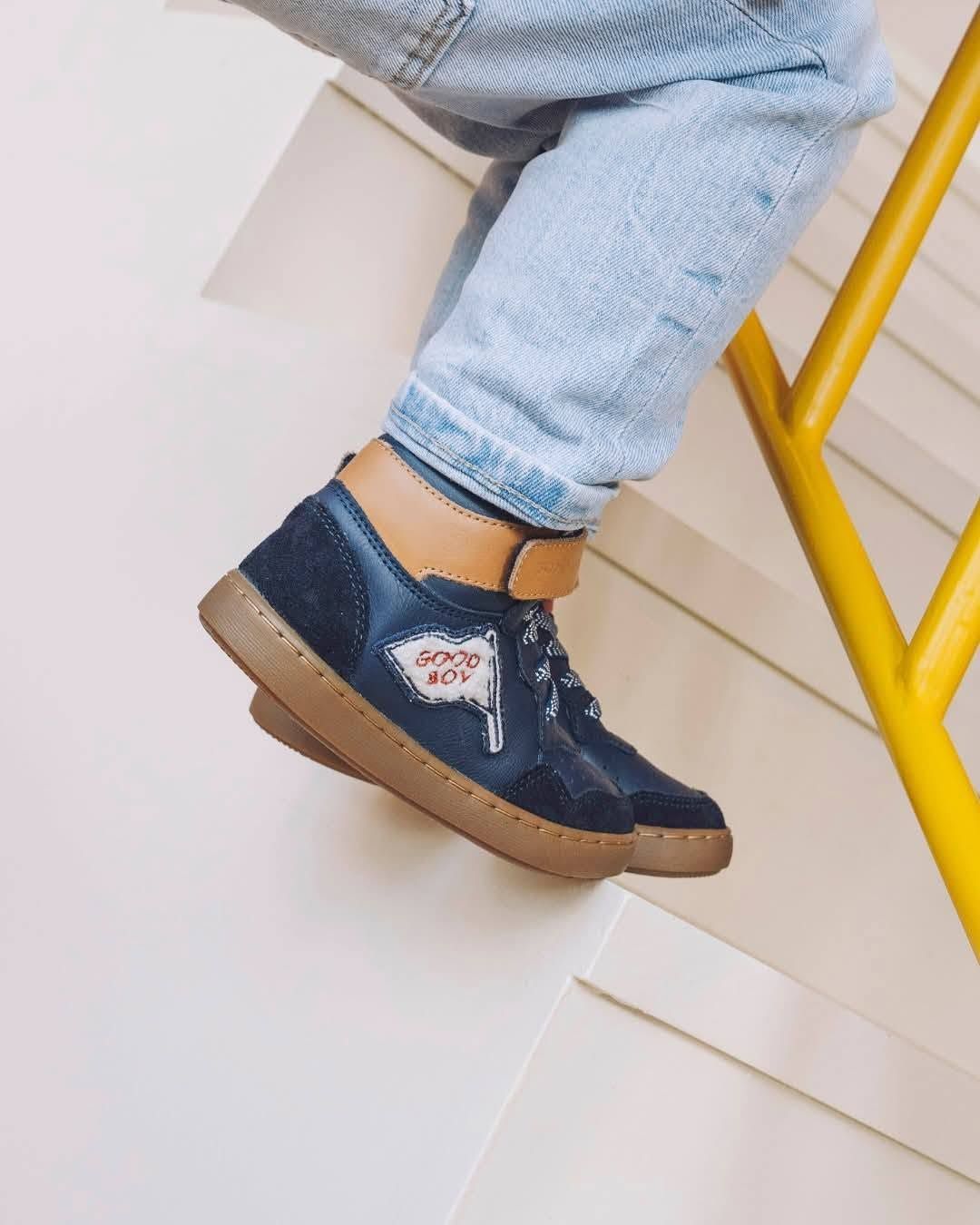Une basket en jean bleu au pied d'une personne, associée à un jean clair, avec une chaise jaune en arrière-plan.