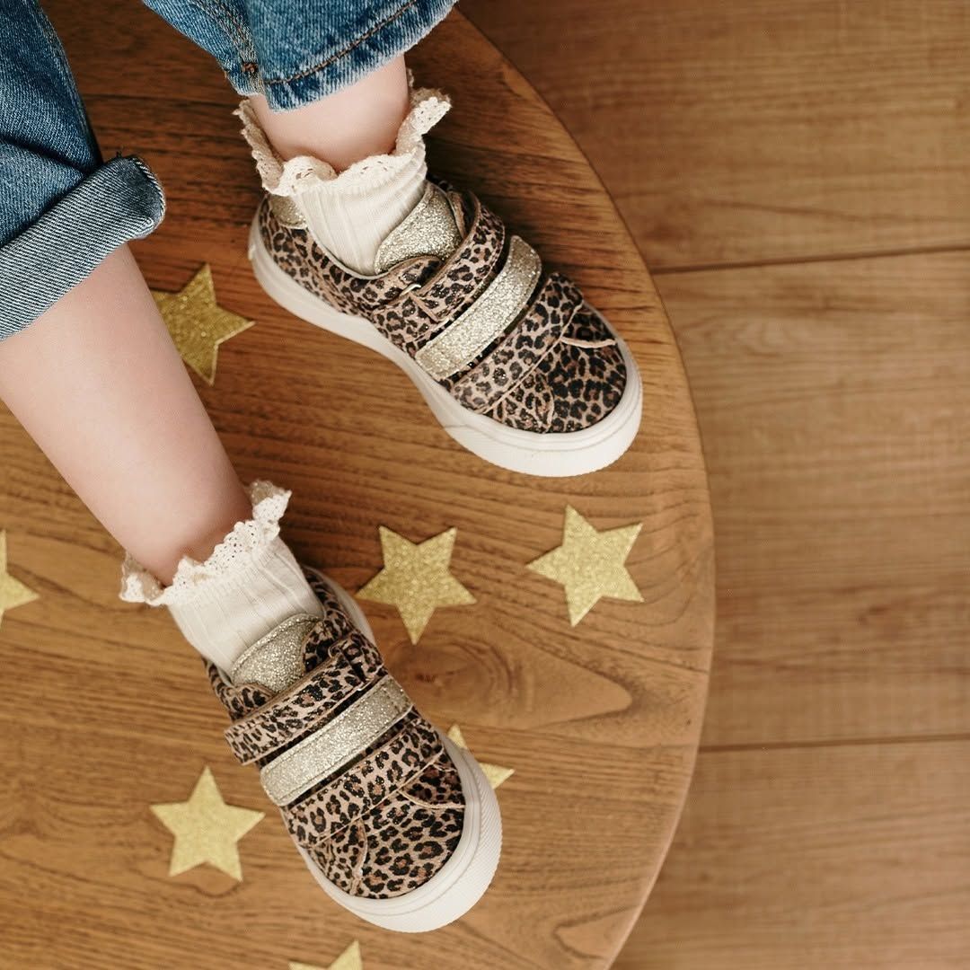 Pieds d'enfant chaussés de baskets à imprimé léopard et de chaussettes à volants, posés sur un tabouret rond en bois découpé en forme d'étoile.