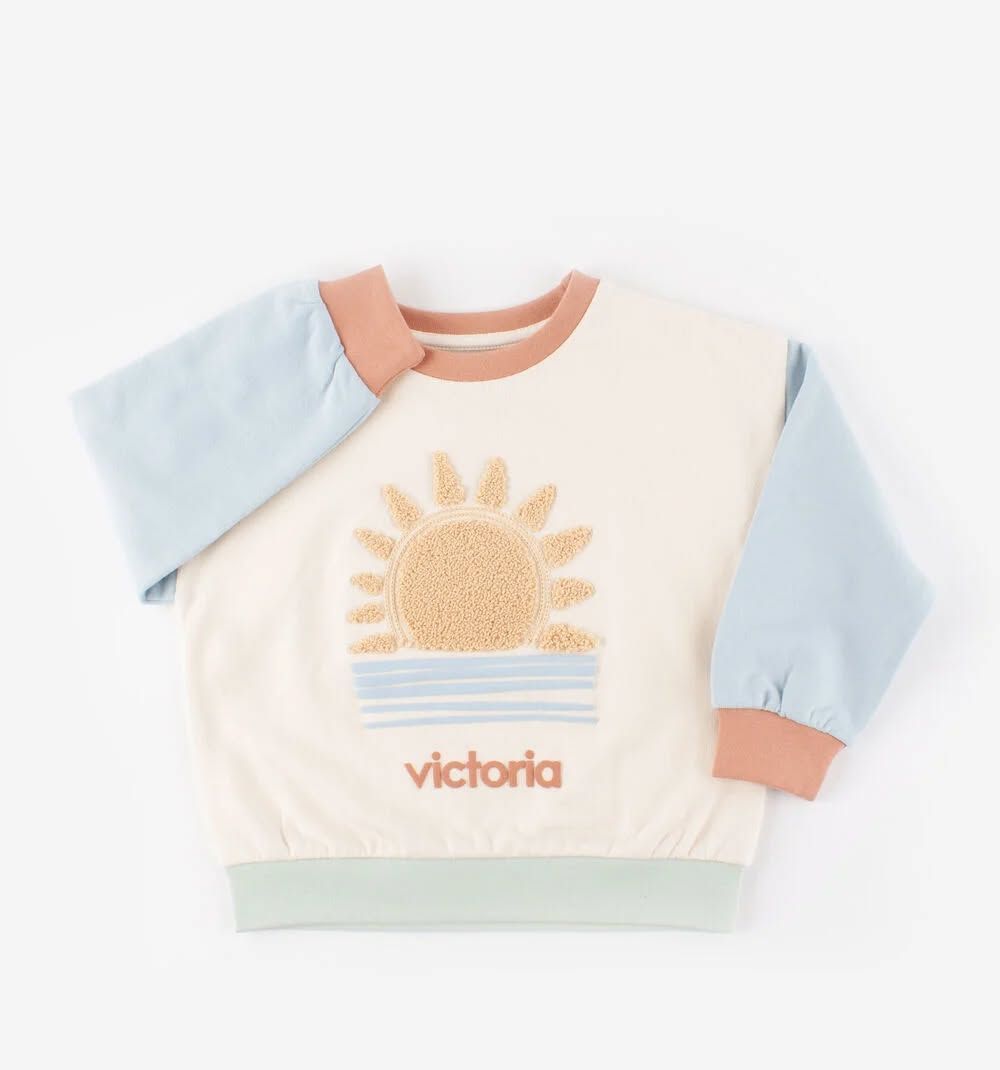 Sweat-shirt bébé pastel avec un motif de soleil et le texte « Victoria » sur fond blanc