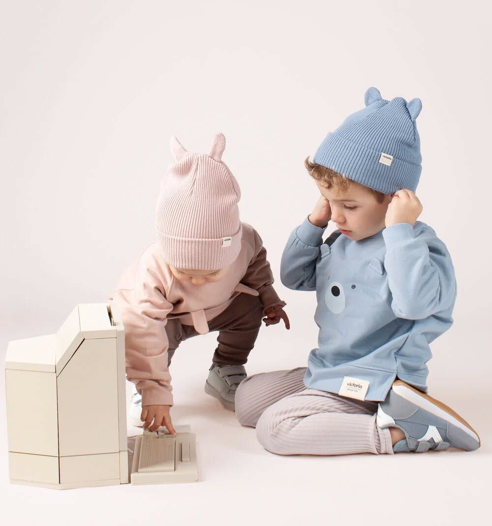 Deux tout-petits portant des bonnets tricotés jouent avec une maison de jouets en bois sur un sol blanc