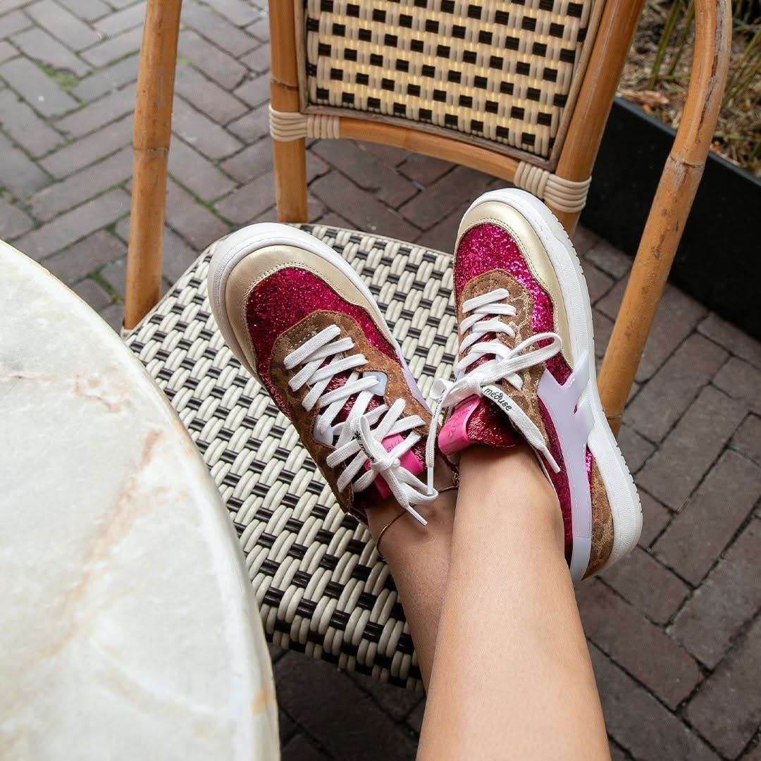 Des pieds chaussés de baskets roses à paillettes reposent sur une chaise en osier près d'une table de café en terrasse.