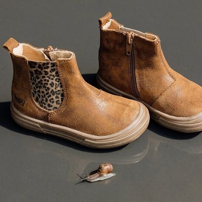 Une paire de bottines marron pour tout-petit, ornées d'empiècements latéraux à imprimé léopard, à côté d'un petit escargot sur une surface sombre.