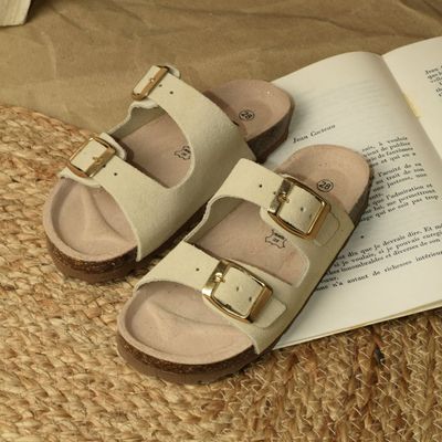 Sandales beiges à double boucle posées sur un tapis tressé à côté d'un livre ouvert