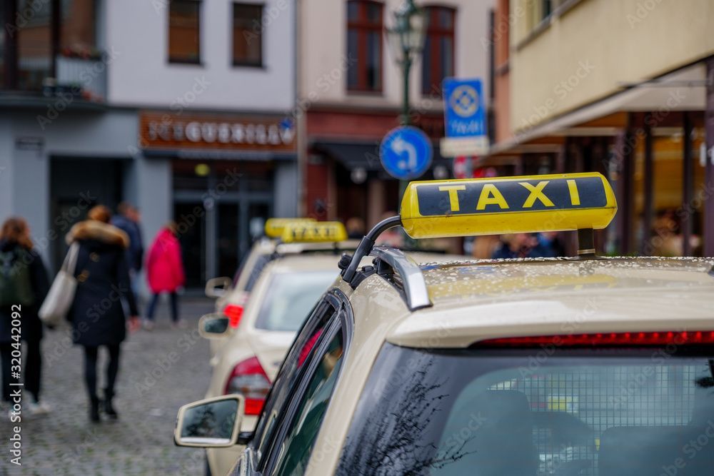 Taxis parken auf einer Kopfsteinpflasterstraße vor Geschäften. Im Hintergrund gehen Menschen spazieren.