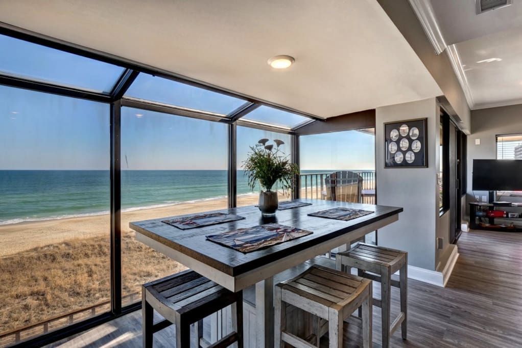 A dining area with beach view. A table with four stools, a vase of flowers, and large windows overlooking the ocean.