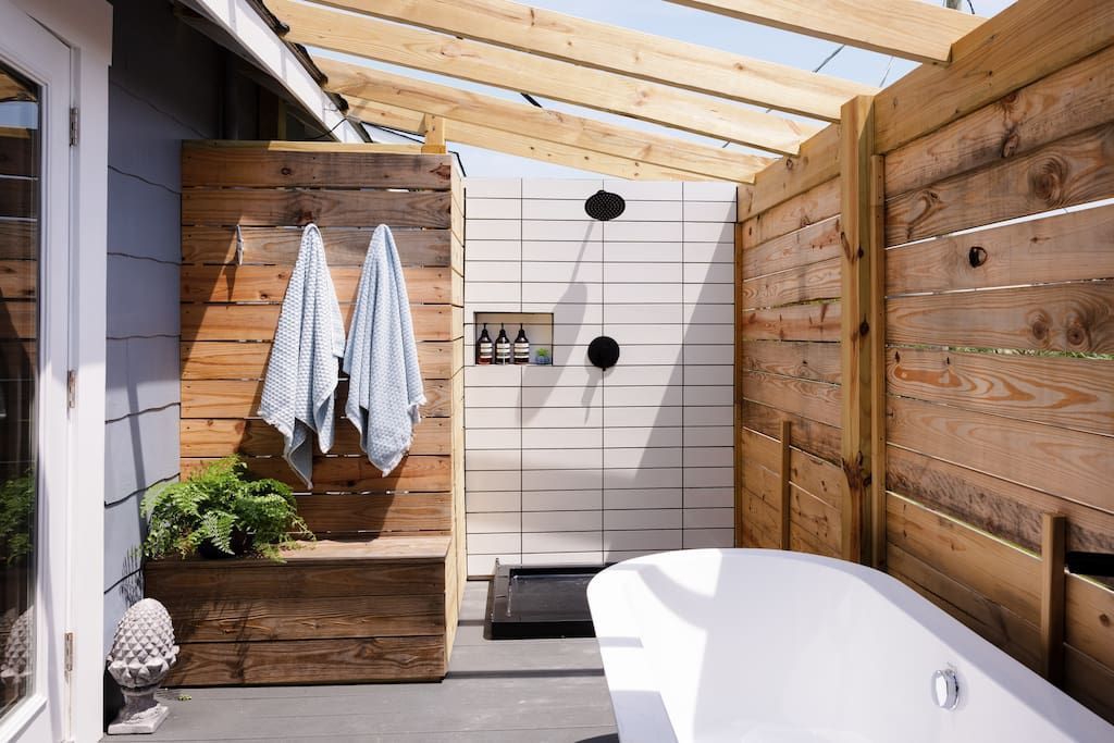 Outdoor shower with wooden walls and tub, light blue towels hanging, light wooden roof.