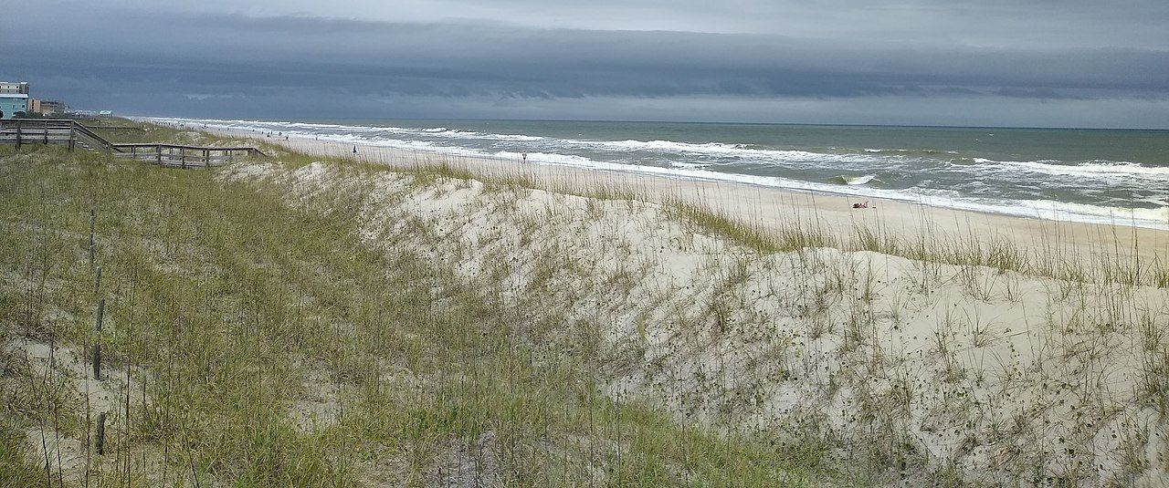 Coastal view of dunes, beach, and ocean under a cloudy sky.