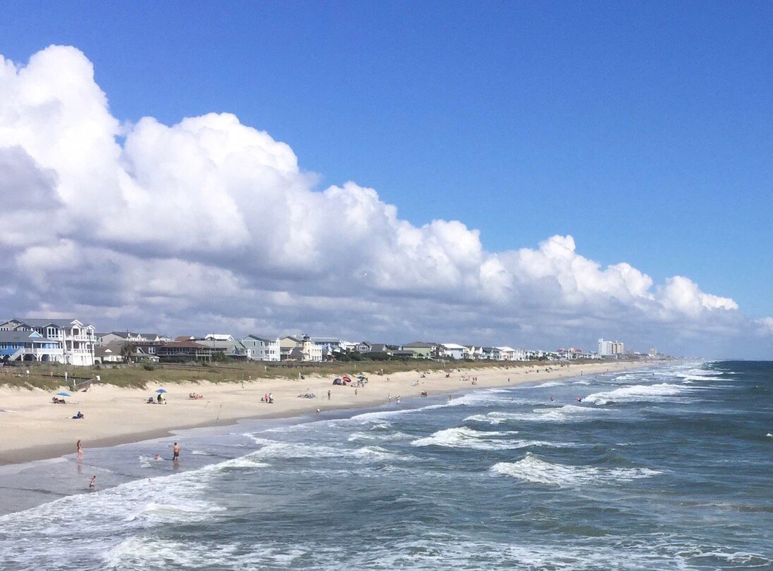 Beach scene with ocean waves, sandy shore, buildings, and blue sky with clouds.