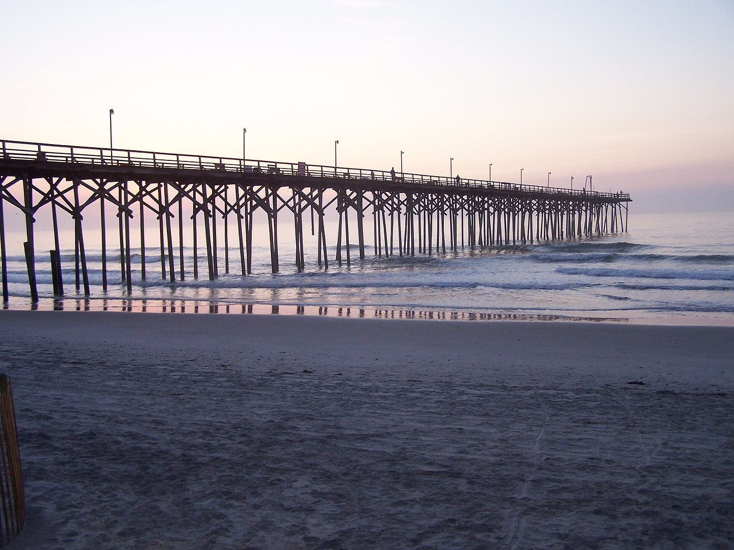 Wooden pier extending over ocean, beach in foreground; twilight sky, calm water.