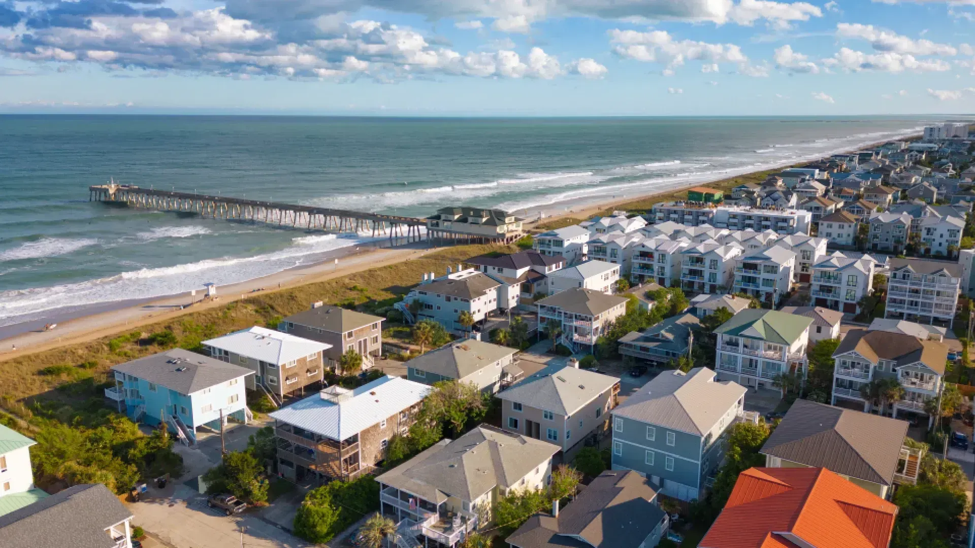 Beachfront houses and a pier along the ocean under a blue sky.