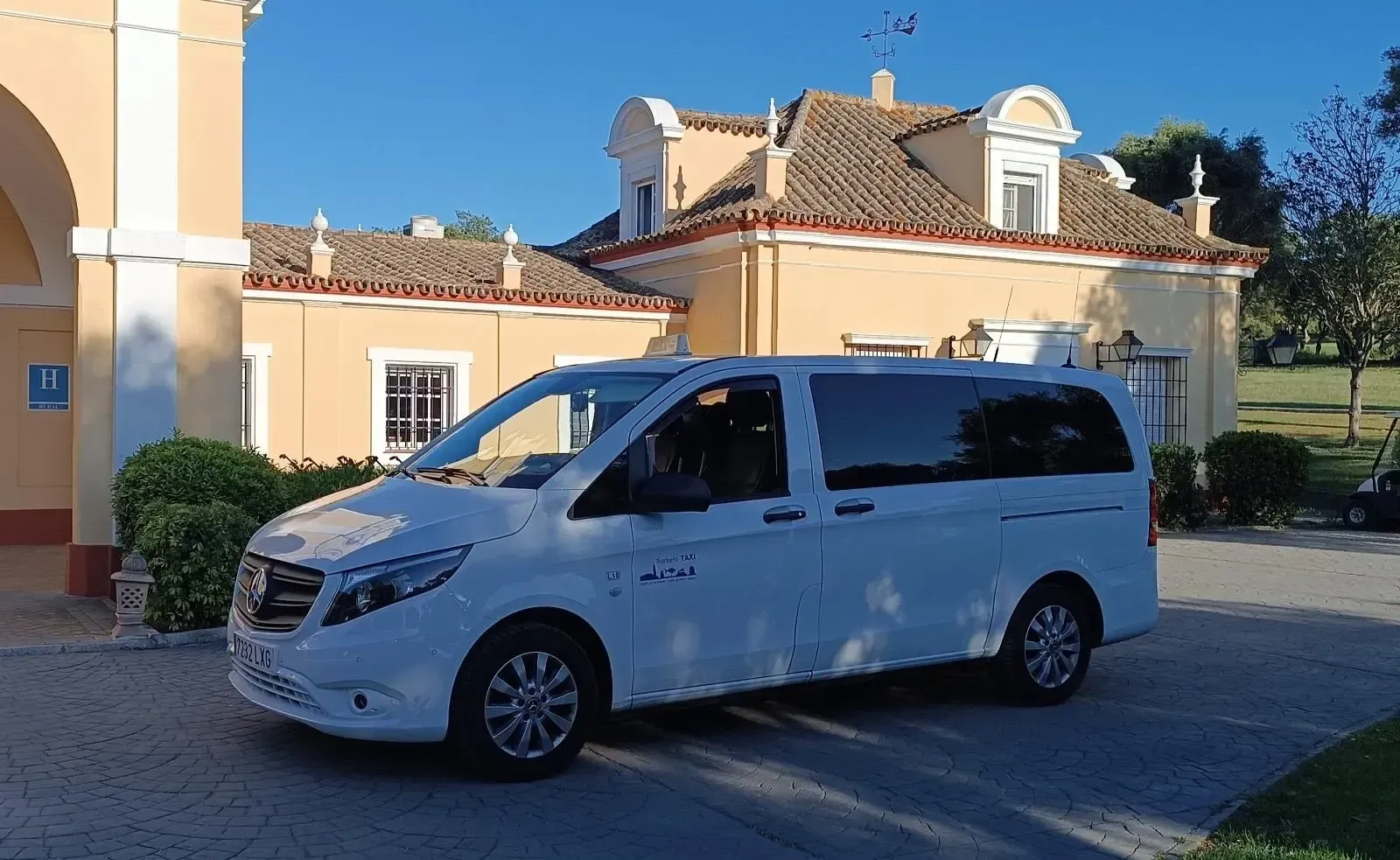 Camioneta blanca estacionada frente a un edificio amarillo claro con techo de tejas rojas y cielo azul.