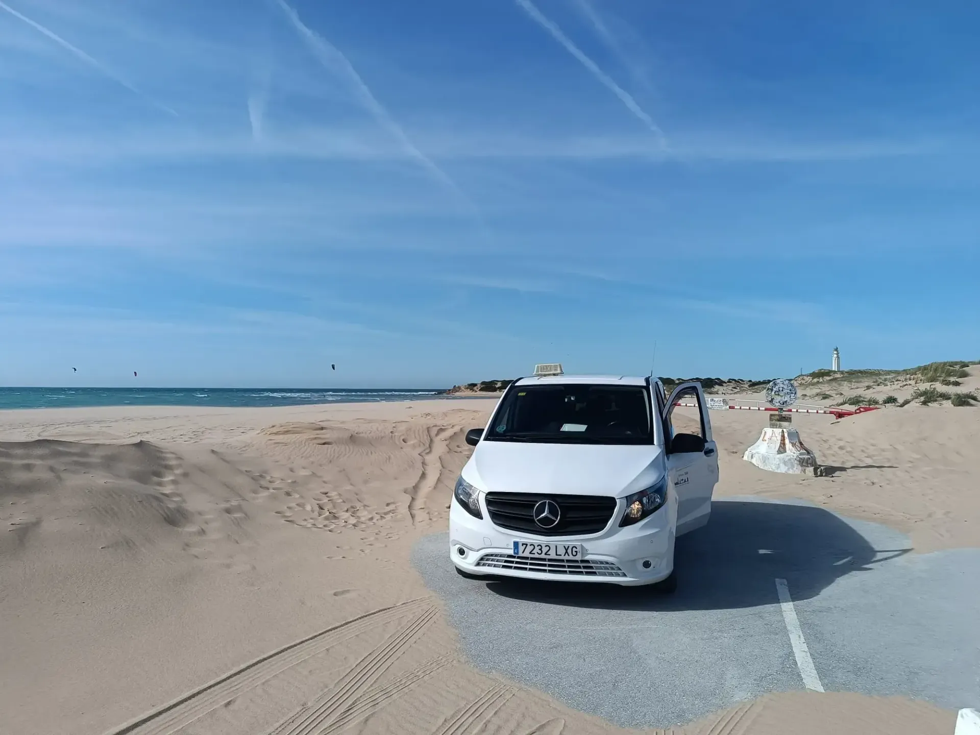 Una furgoneta Mercedes blanca estacionada en una playa de arena bajo un cielo azul. El océano se ve al fondo.