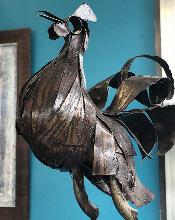 Sculpture de coq en métal sur un mur bleu ; plumes texturées et détails soignés