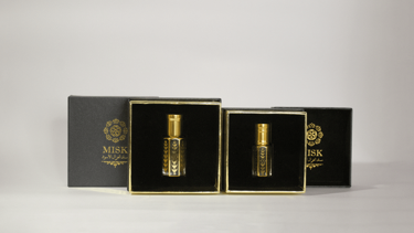 Dos cajas de perfume negras y doradas con pequeños frascos dorados expuestos sobre una superficie blanca.
