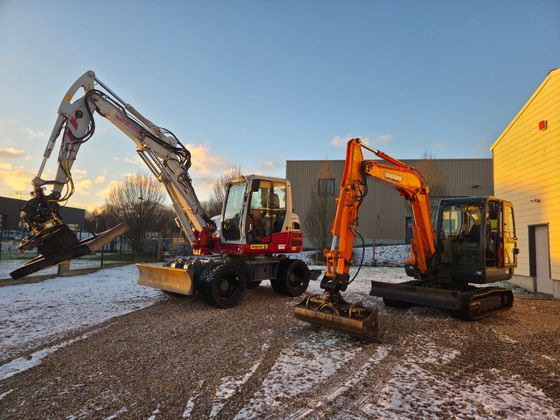 Deux excavatrices, une rouge et une orange, travaillent sur un chantier enneigé au coucher du soleil.
